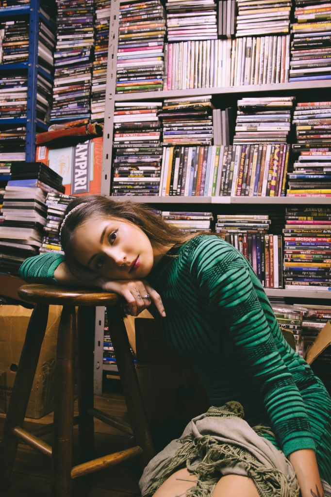 Bárbara Luiza encostada em um banco de madeira, em frente a uma estante repleta de CDs e DVDs